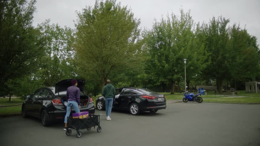 Two people are at a park taking boxes out of the trunk of their car and putting them into a wagon when another car pulls in and the man joins them