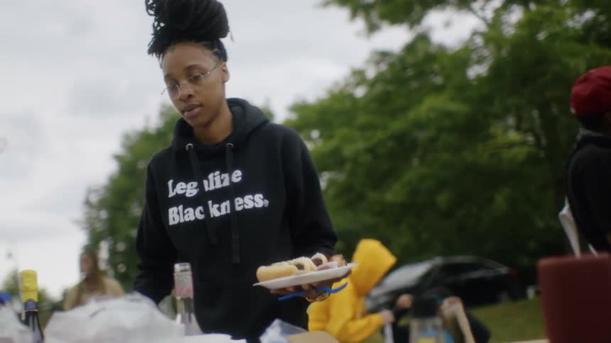 Woman puts together plate with hotdogs as friends and family talk behind her at picnic in the park