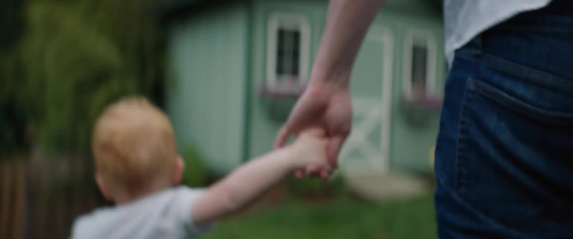 A man is holding his young sons hand as they walk through the yard to the play house