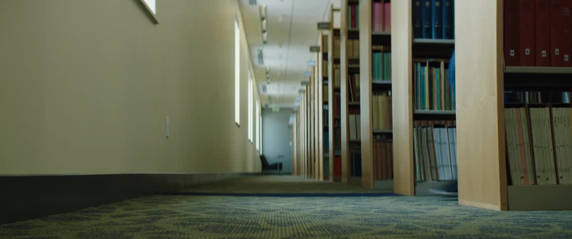 There are rows of book shelves in a library and someone comes out of an aisle and walks away