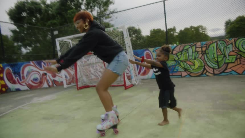 Child pushes mom on roller skates as family and friends mill around graffiti mural decorated concrete soccer field