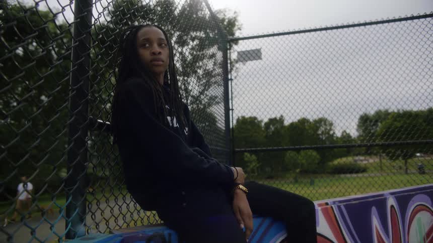 A teenager is sitting on a fence and leaning against the chain linked fence at a park