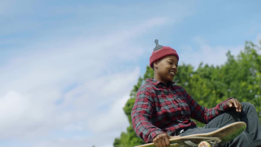 A teenager wearing a knit cap and a flannel shirt is carrying a skateboard and she leaps onto a concrete barrier and sits down smiling