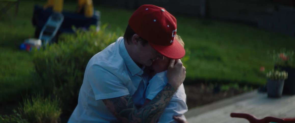 A man wearing a red cap is outside hugging his young son