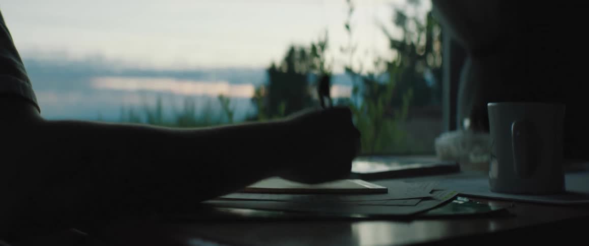 A man sitting at the table in front of a large window writing and thinking
