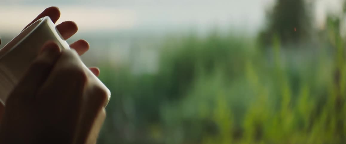 A woman drinks coffee while standing next to her window
