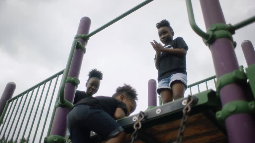 A young girl is climbing up a wall in a playground and two girls are on top clapping and cheering for her when she gets to the top