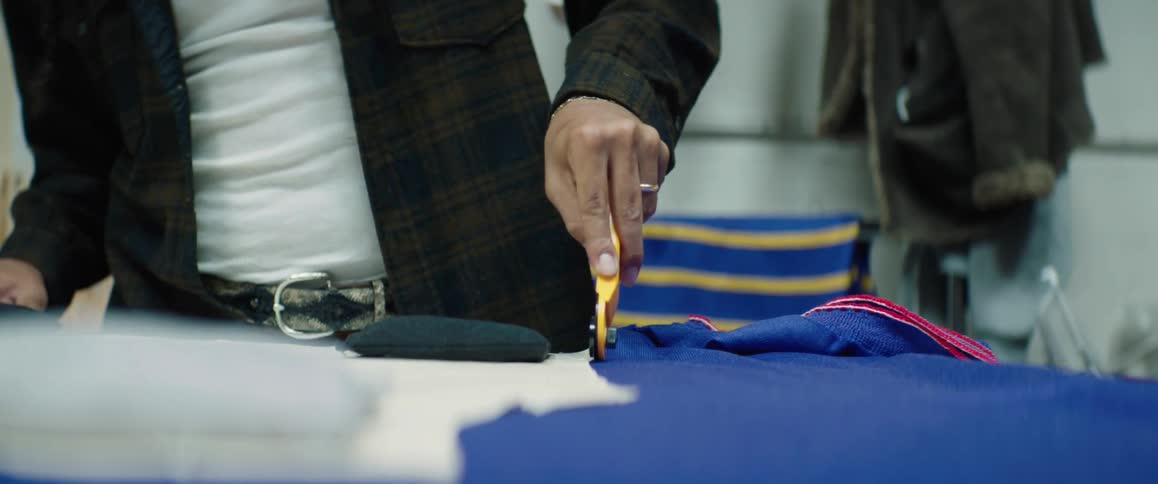 Male tailor with white and dark green shirt uses rotary cutter cutting blue cloth