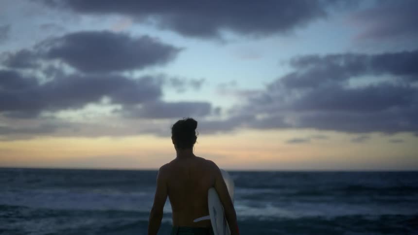 A man standing on the beach holding his surfboard looking out at the waves and ocean