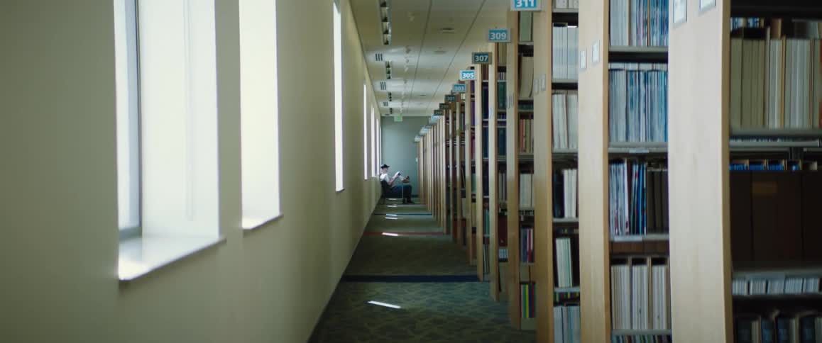 There are rows of book shelves in a library and a man sitting at the end of the aisle gets up and walks away