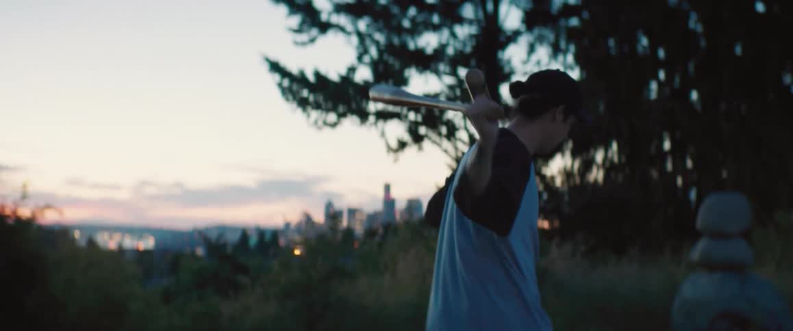 Man with cap and gray shirt sways basball bats walking in the field at dusk