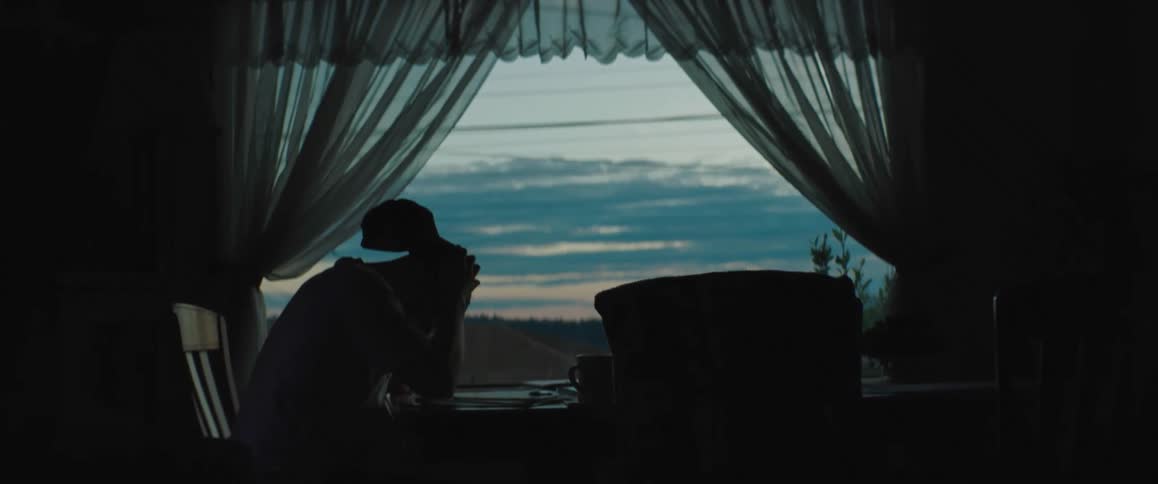 A man is sitting at a table looking out the window and he takes off his cap and scratches his head