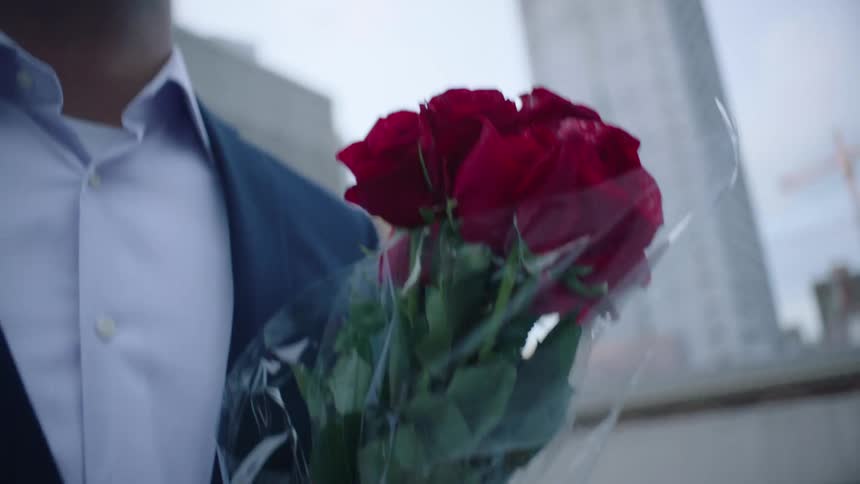 A man is walking on a downtown street carrying a bouquet of roses and there are tall buildings in the distance