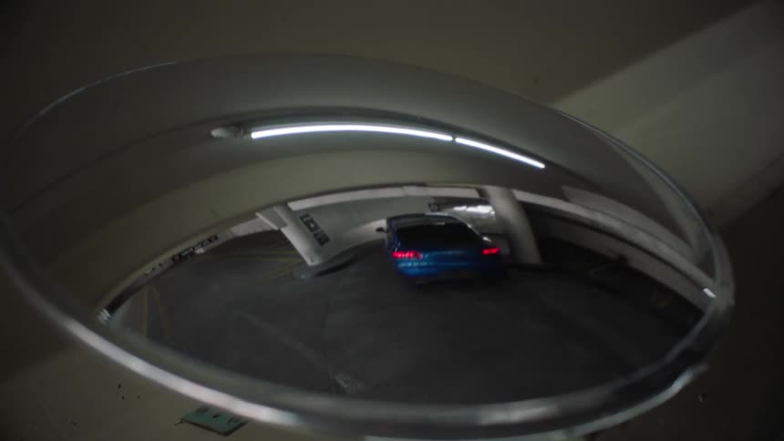 A convex mirror in a parking garage reflects a blue car traveling down a ramp
