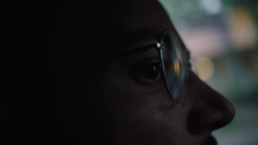 A man wearing glasses is traveling in an automobile with buildings and lights passing by in the window