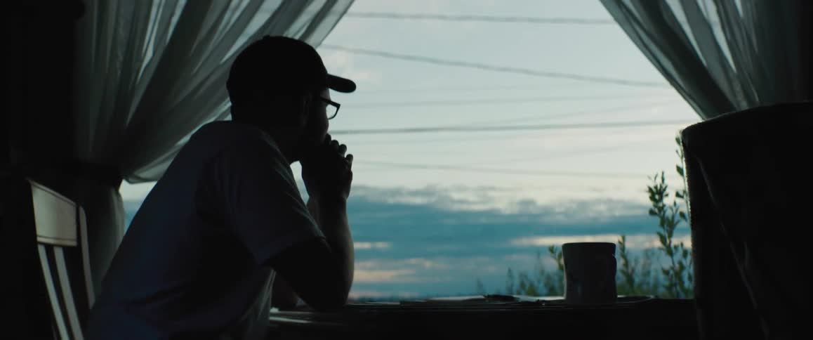 The silhouette of a man sitting at a table looking out the window removing his cap and hanging his head in despair