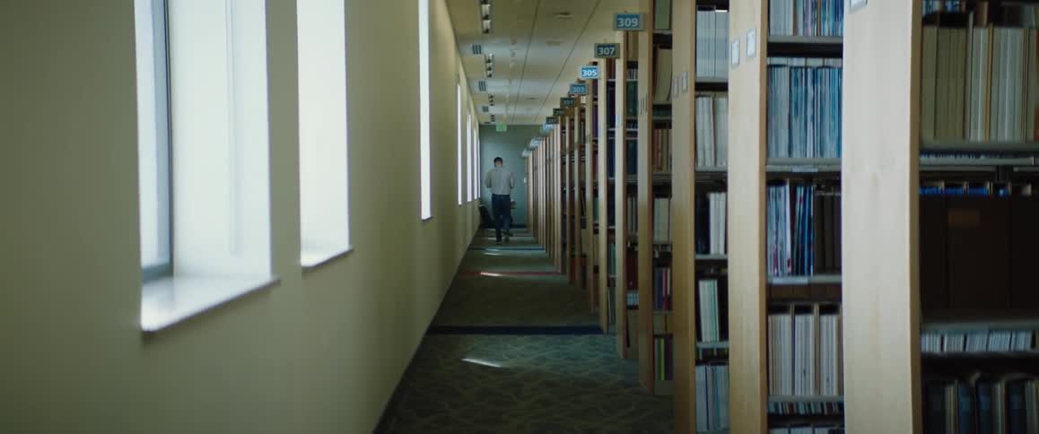 Man with white shir and jeans walks towards desk and chairs and sits down in the large library