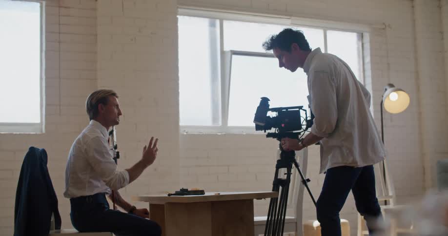 A cameraman films and instructs a man sitting down in a suit
