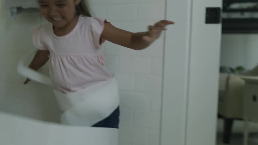 A little girl is laughing as she plays in a bathroom wrapping herself in toilet paper
