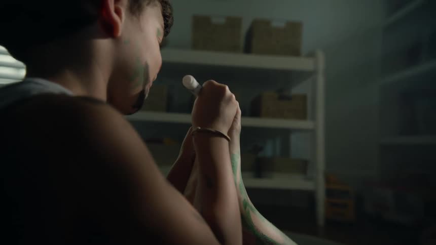 A little boy with marker ink all over his face is sitting on the floor coloring on another boys foot with a marking pen