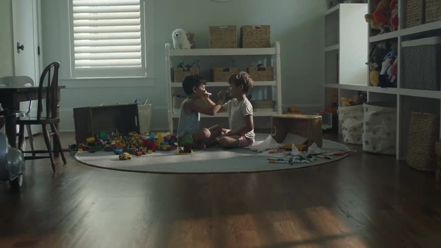 Two little boys sitting on a playroom rug drawing on each others faces with marker