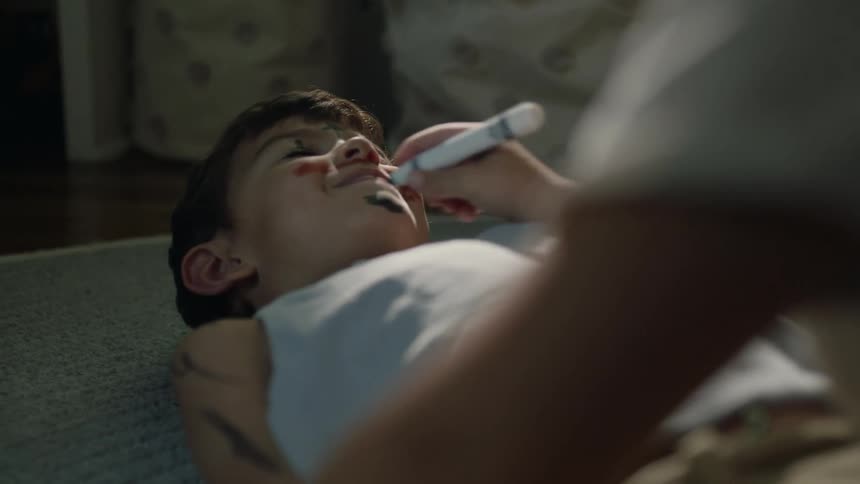 A boy is lying on the floor and another boy is coloring his face with a marking pen
