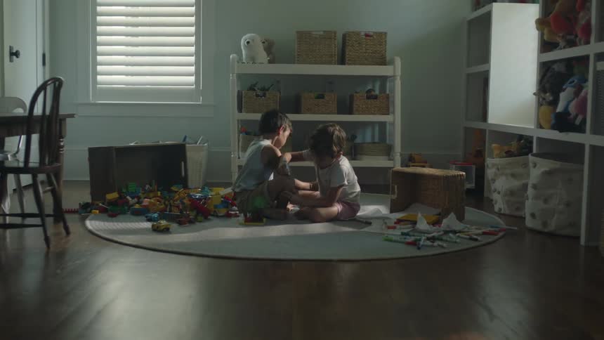Two little boys are sitting on the floor in a room with toys writing on each other with marking pens