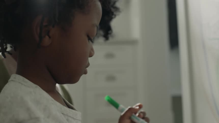 A teenage girl is holding a young girl in her lap as she draws on the wall with colored marking pens