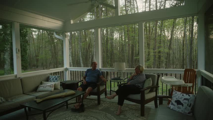 Two people are sitting in an enclosed patio with a ceiling fan spinning overhead and woods are outside