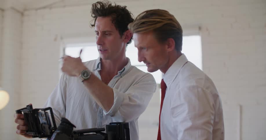 Two filmmakers standing in front of a camera talk while setting up a shot inside a white room