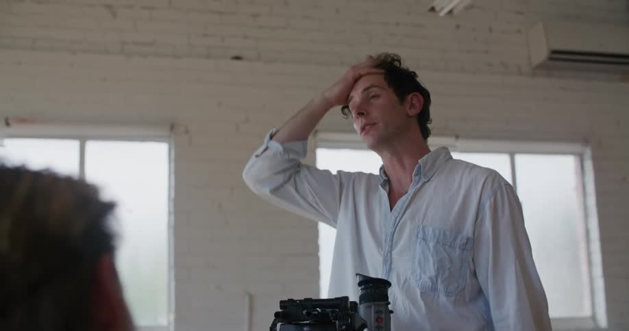 A man directing an interview talks and gestures with his hands before leaning down to look through the film camera