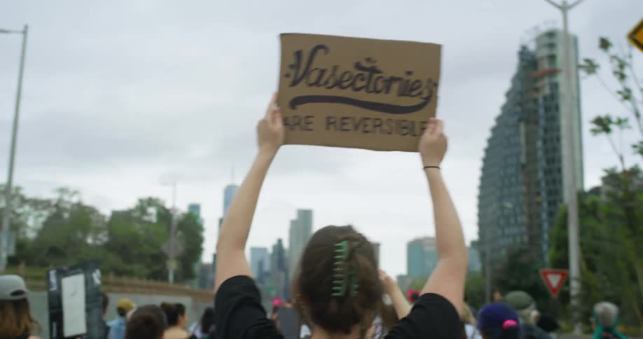A woman holds up a cardboard sign while marching through a city with a crowd of protesters