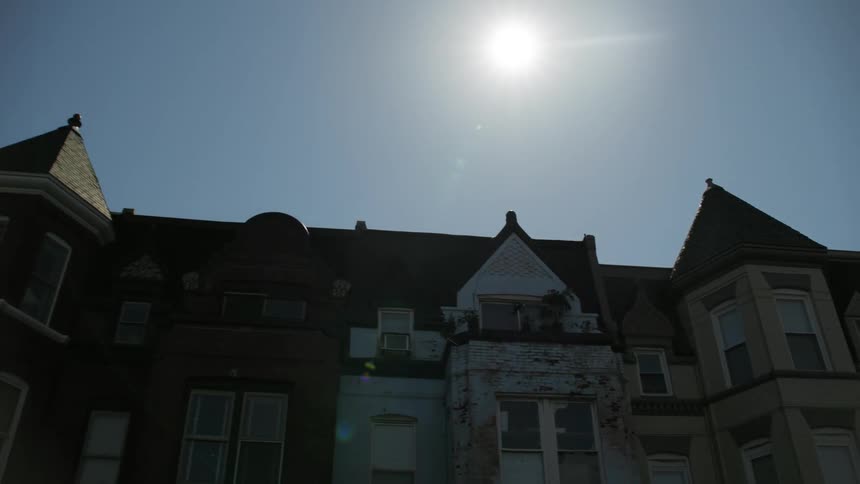 A bright sun in the blue sky above row houses on a city street
