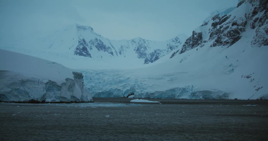 Pieces of ice are floating in the water near snow covered mountains with peaks high in a cloudy sky