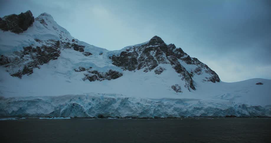 Pieces of ice are floating in the water near snow covered mountains with peaks high in a cloudy sky