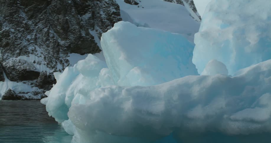 A melting iceberg is floating on the water and snow covered mountains are in the distance