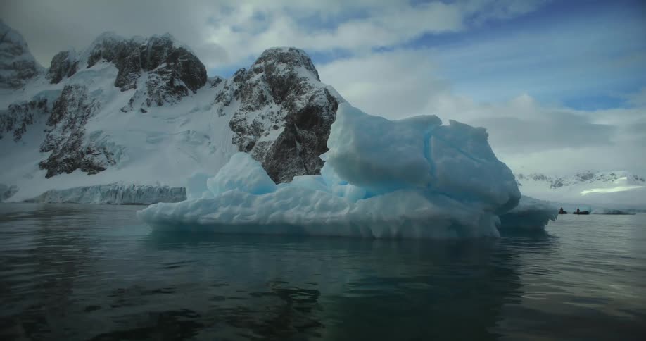A melting iceberg is floating in the water and snow covered mountains are in the distance