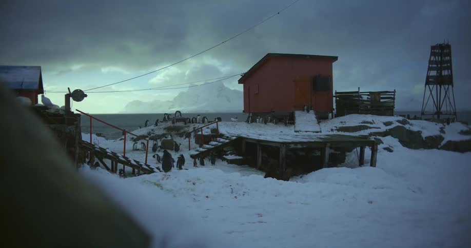 Snow is on the roof of an old building and there are barrels near a fence under a cloudy sky