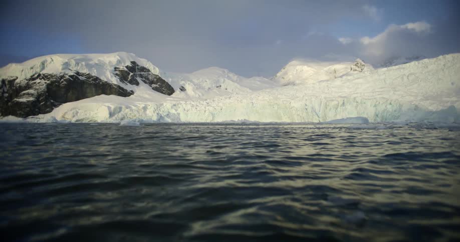 Pieces of ice are moving in the water as sunlight reflects on the ripples and waves and there are snow covered mountains in the distance