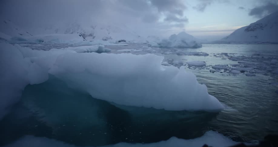 A melting iceberg is floating on the water and snow covered mountains are in the distance under a cloudy sky
