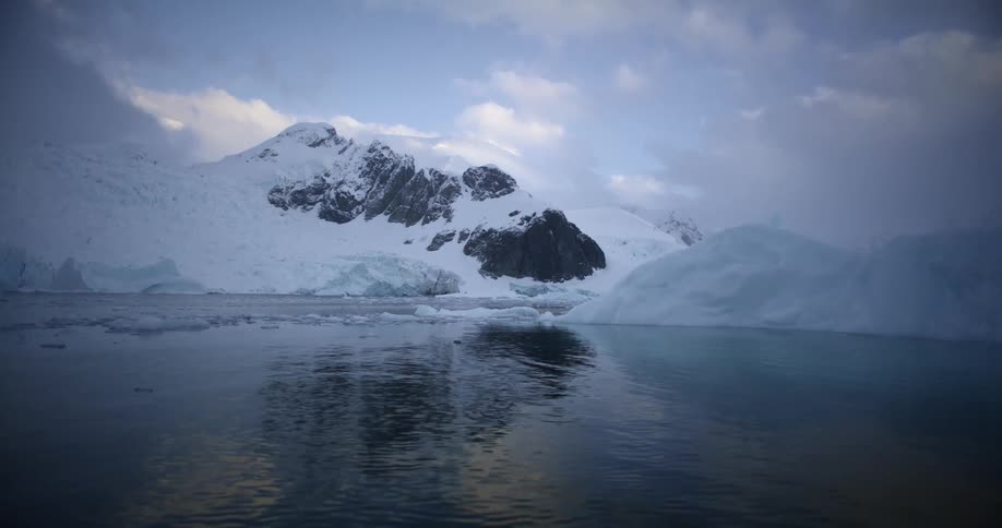 Snow covered pieces of melting ice are floating in the water with snow covered mountains in the distance under a cloudy sky