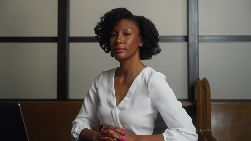 A woman sits with her hands crossed in front of her and turns her head and looks ahead