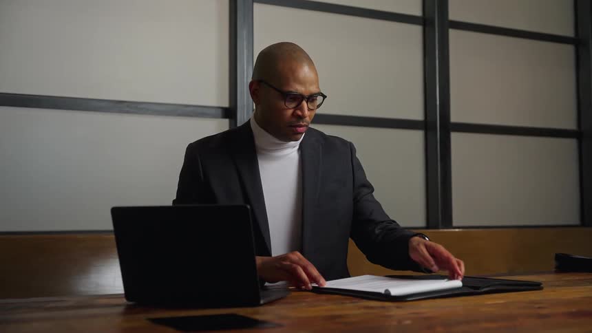 A man sitting at a table in an office opens a laptop and prepares his papers and pen