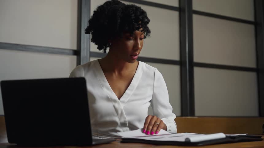 Woman sits at desk using laptop and going through paperwork