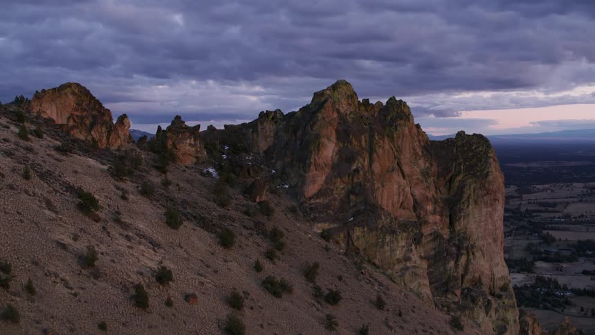 Plants grow on a mountain ridge overlooking the land below and mountains are on the horizon under a cloudy sky