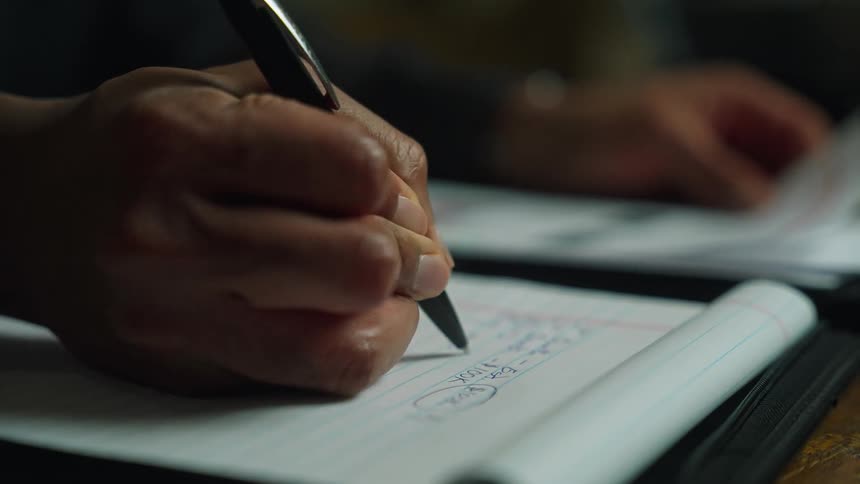 Someone is sitting at a table holding a paper in their left hand and writing on a notepad with their right hand
