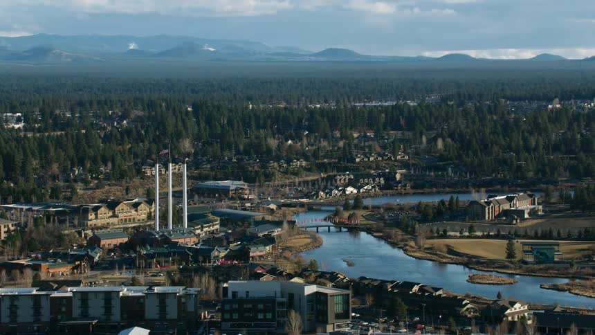 There is a community along a river and automobiles are traveling over a bridge with forests and mountains in the distance under a cloudy sky