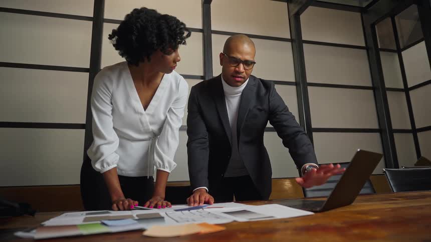 A man and woman discuss paperwork on a laptop in a conference room