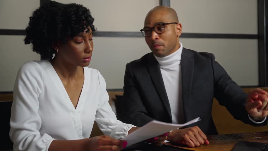 A man and a woman sit in an office and go through papers and look at a laptop