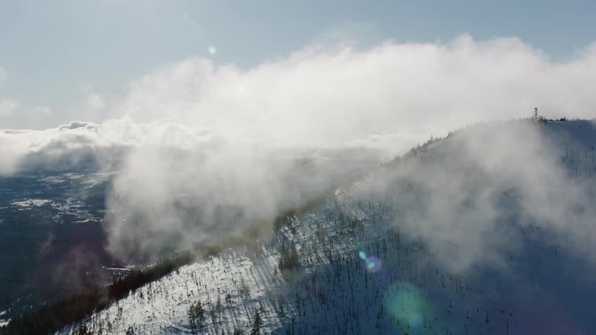 The sun shines upon a snow covered forested mountain high in the clouds